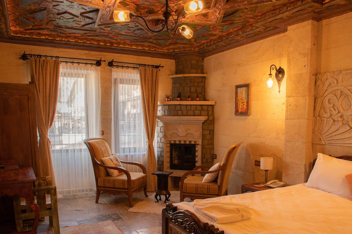 A hotel room with a fireplace, two chairs, a bed, and a nightstand with a lamp and phone.