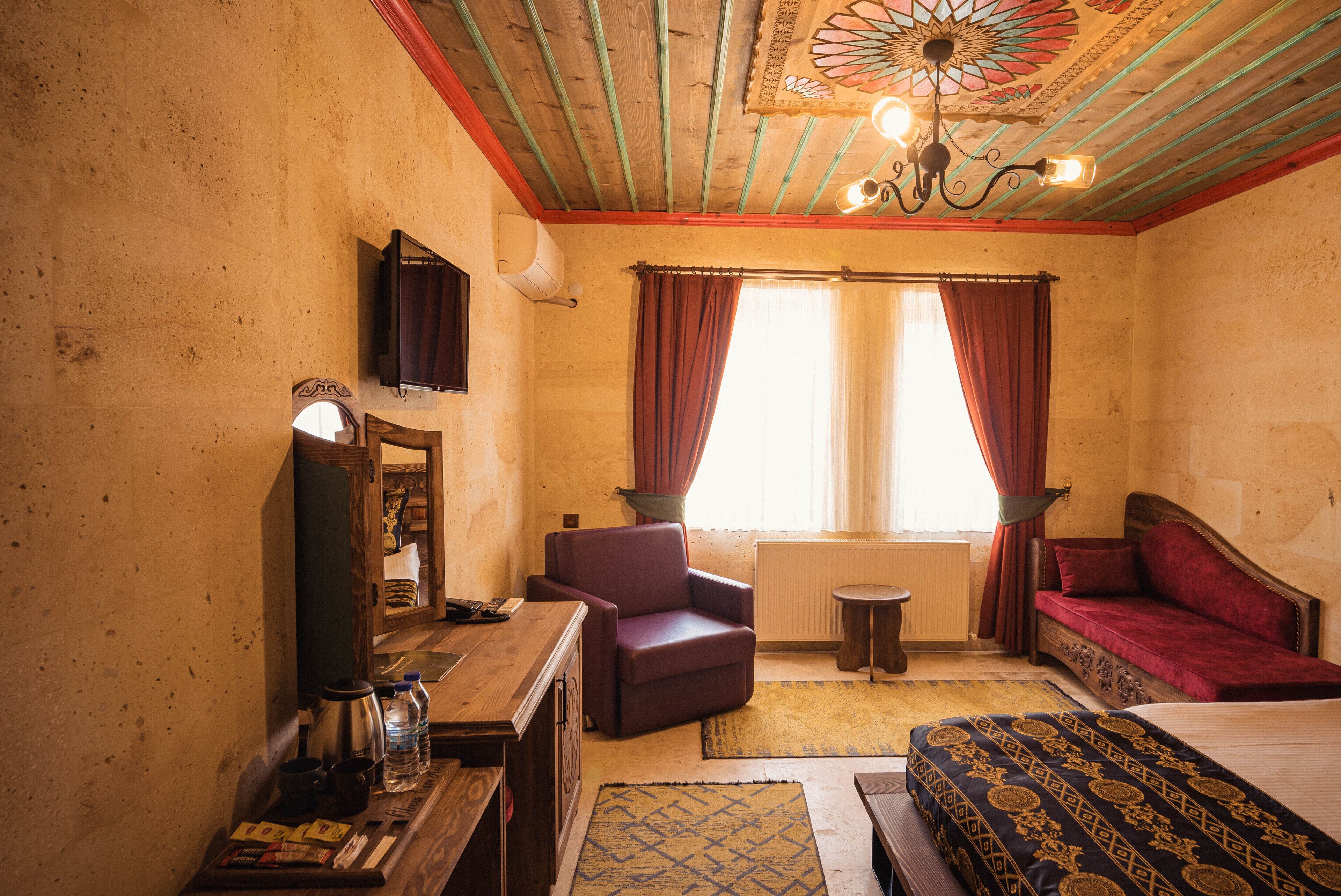 A room with a wooden ceiling, a bed with a patterned bedspread, a red sofa, a purple armchair, a small table with a lamp, and a window with curtains.