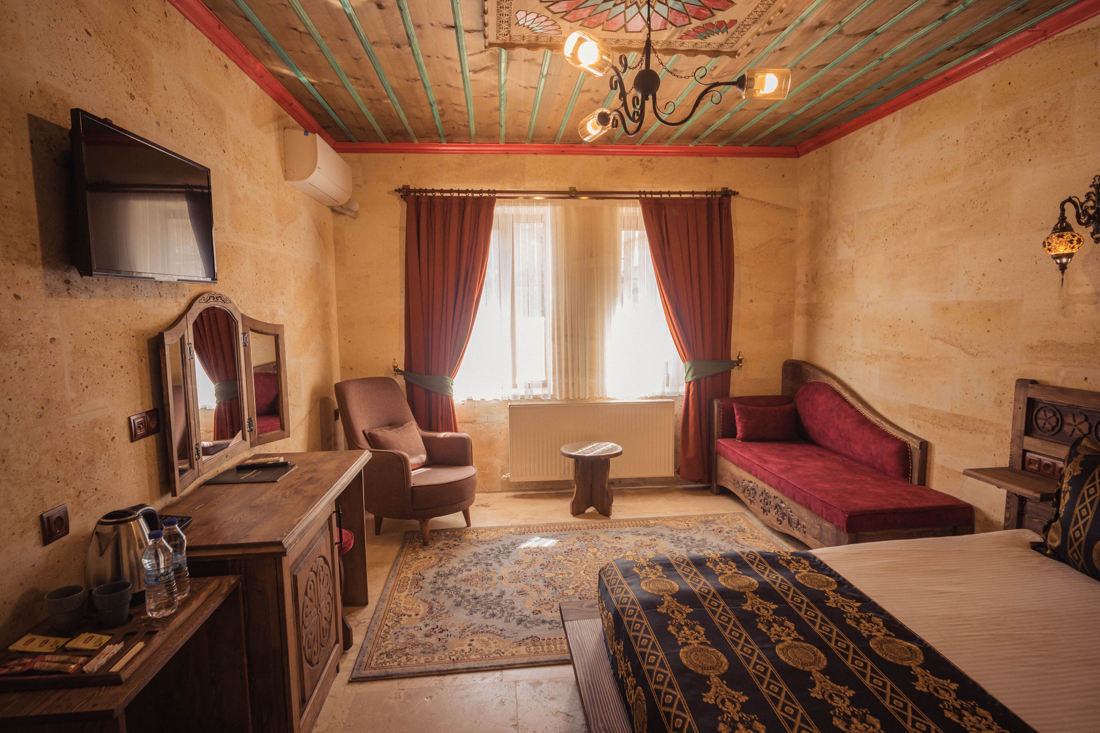 A room with a bed, a chair, a sofa, a small table, a mirror, a radiator, a television, and a window with curtains.