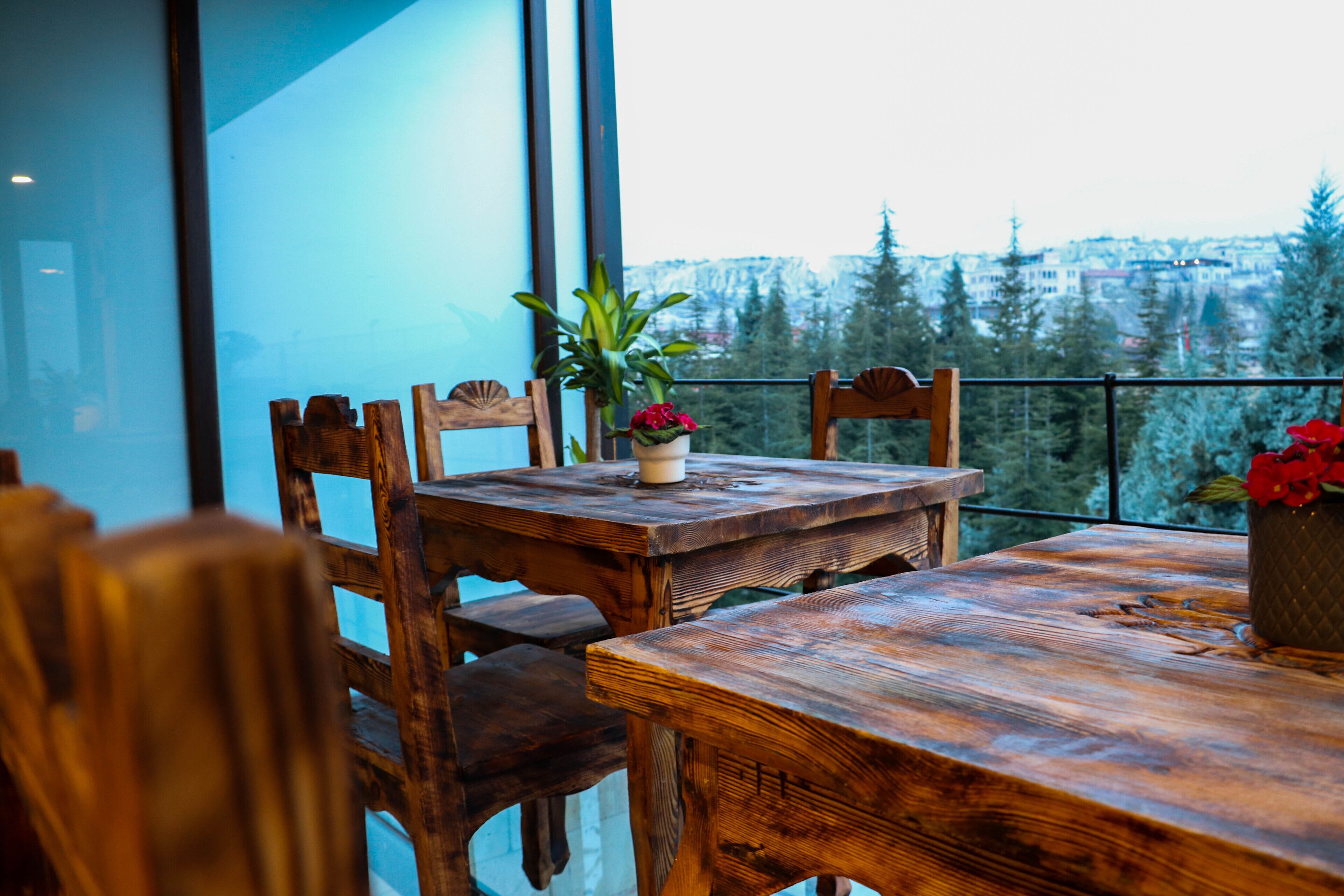 Une table à manger en bois, entourée de chaises, un pot de fleurs et une vue sur le paysage urbain à travers de grandes fenêtres.