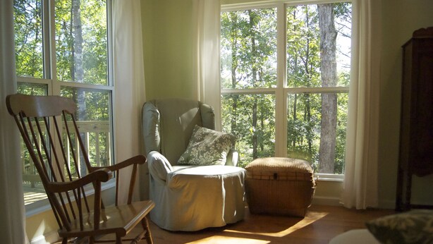 A room with a wooden rocking chair, a cushioned chair, and a small ottoman near large windows with a view of trees. - Scout's Hideaway on Beautiful Smith Lake (Arley)