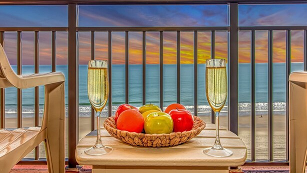Un balcon avec vue sur l’océan, deux coupes de champagne et un panier de pommes aux couleurs vives.