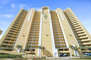 A high-rise hotel with multiple balconies, a prominent logo, and a parking area with a few cars.