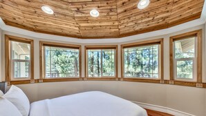 Une chambre avec un grand lit, un plafond en bois et de nombreuses fenêtres donnant sur la forêt.