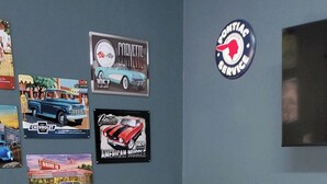 Un petit coin repas avec une table ronde et deux chaises, un mur décoré d’affiches de voitures anciennes et une enseigne Texaco.