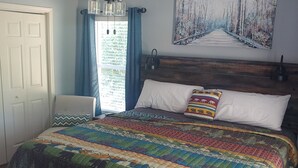 Une chambre avec un lit en bois, un ventilateur de plafond, un tableau représentant une forêt et une fenêtre avec des rideaux bleus.