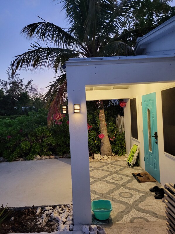 A small, white building with a turquoise door, a tiled patio, and a palm tree in the background.