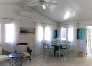 A bright, airy room with a ceiling fan, a dining table with chairs, a sofa, and two windows with curtains.