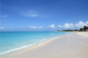A beach with clear turquoise water, sandy shore, and a few buildings in the background.
