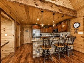 A rustic kitchen with wooden cabinets, a stainless steel refrigerator, a stone island with bar stools, and pendant lighting. - Blue Ridge Mountaintop Cabin with a Stunning View, Hot Tub, Pool Table, Fire Pit (Blue Ridge)