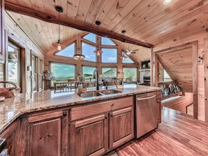 A spacious kitchen with a granite countertop, wooden cabinets, and a large island. There are multiple windows offering views of the surrounding landscape.