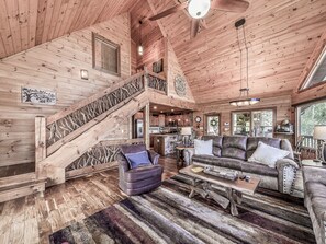 A spacious log cabin interior with a wooden staircase, a cozy living area with leather sofas, a dining table, and a kitchen visible through an open door.