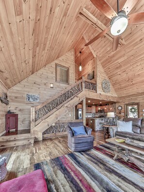 A spacious log cabin interior with a high ceiling, a wooden staircase, a cozy living area with a leather sofa, a dining table, and a kitchen in the background.