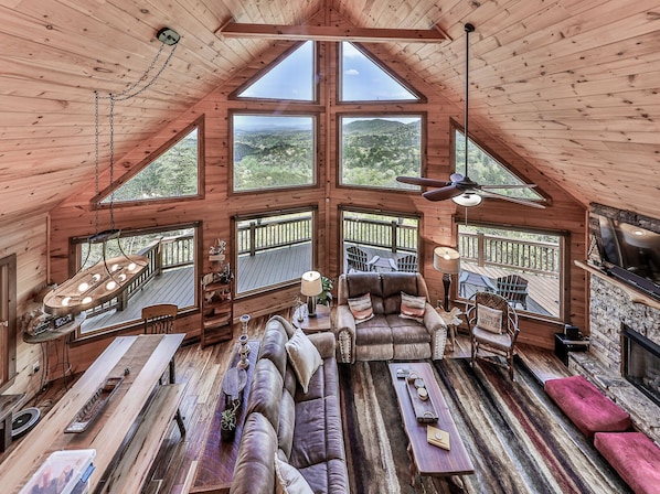 A spacious loft-style room with a high ceiling, large windows offering views of the outdoors, a cozy seating area with a sofa and chairs, and a dining table with chairs.