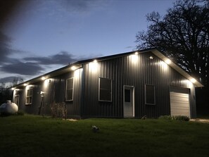 A metal building with multiple windows and a garage door, illuminated by exterior lights. - The "Shouse"- Take the road less traveled- for fun & relaxation in the country (Ossian)
