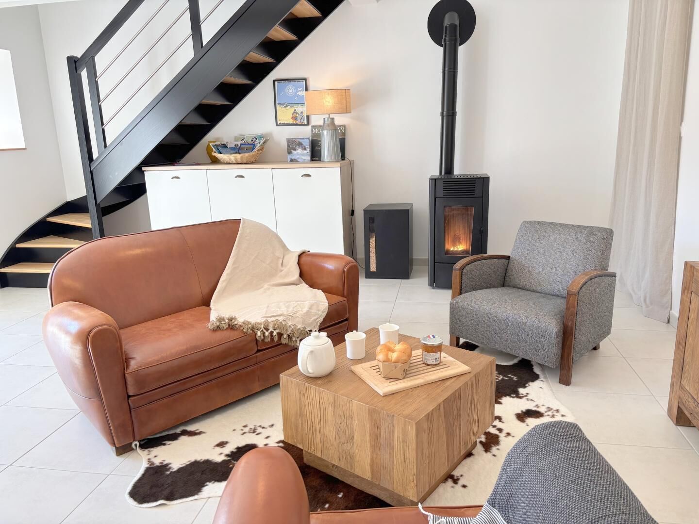 A modern living room with a brown leather sofa, a grey armchair, a wooden coffee table with a cowhide rug, a fireplace, and a staircase.