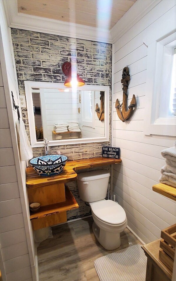 A bathroom with a rustic brick wall, a wooden vanity, a white toilet, and a decorative anchor on the wall. - Welcome to Sunshine Shores. Just steps from the condo door to PCB sandy shores. (Panama City Beach)