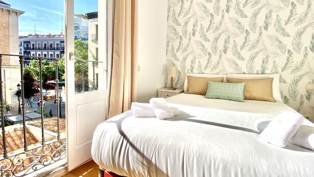 A hotel room with a bed, a nightstand, a window with curtains, and a view of the cityscape. - Huertas II - 2 bedrooms, in the best area (Madrid)