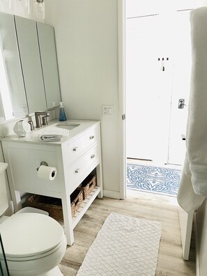 Une salle de bain avec un lavabo blanc, un miroir et des toilettes. Il y a un tapis blanc sur le sol et une porte qui donne sur l’extérieur.