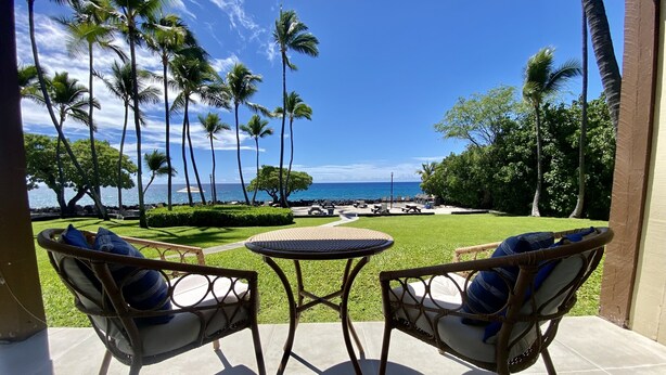 Eine Terrasse mit Korbstühlen und einem kleinen Tisch, mit Blick auf einen Strand mit Palmen und dem Meer.
