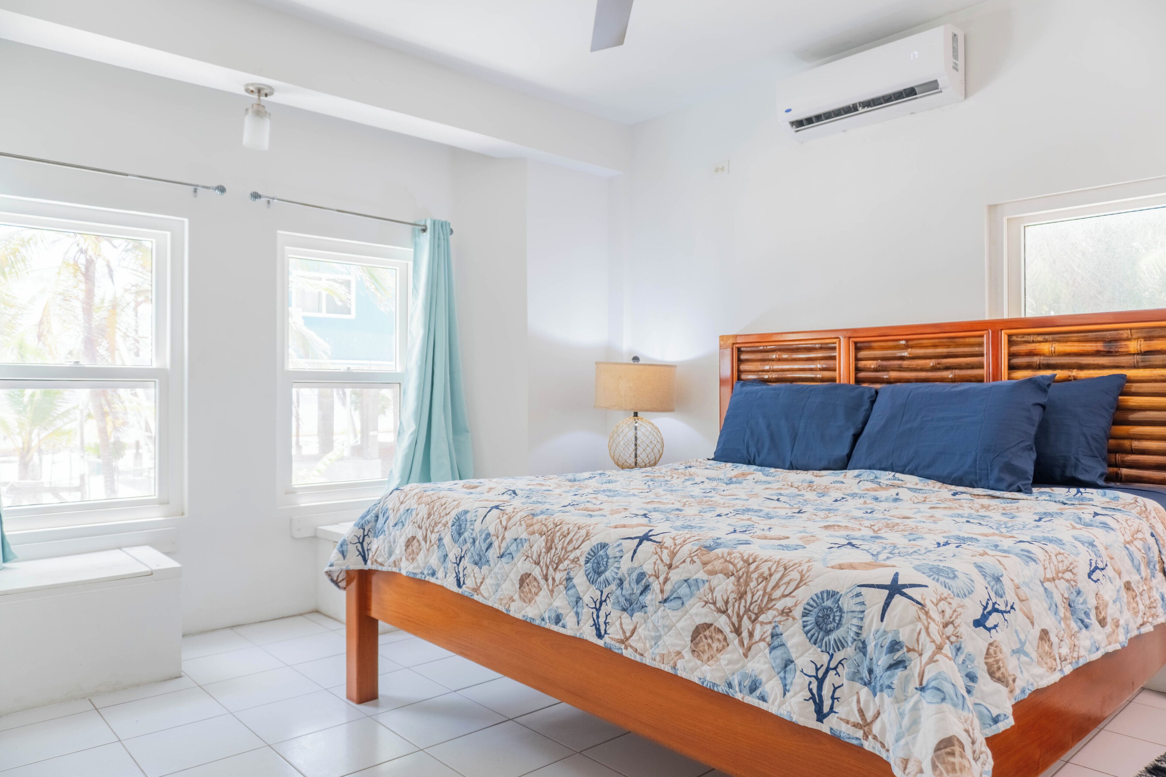 A bedroom with a bed, a window, a wall-mounted air conditioner, and a bedside lamp.