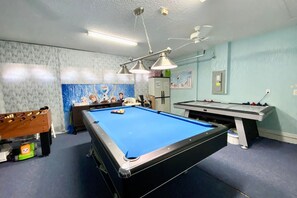 Une table de billard recouverte d’un drap bleu, une table de baby-foot et une table de jeu de palet dans une pièce aux murs bleu clair et dotée d’un ventilateur de plafond.