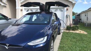 Une Tesla Model 3 est garée devant un garage résidentiel, dont les portes sont ouvertes.