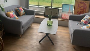 Ein Zimmer mit grauem Sofa, einem kleinen Tisch mit grüner Vase und einem großen Fenster mit Blick auf ein grünes Feld.