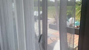 Ein Zimmer mit transparenten weißen Vorhängen, Holzboden und Blick auf eine Terrasse mit Pool und Pflanzen.