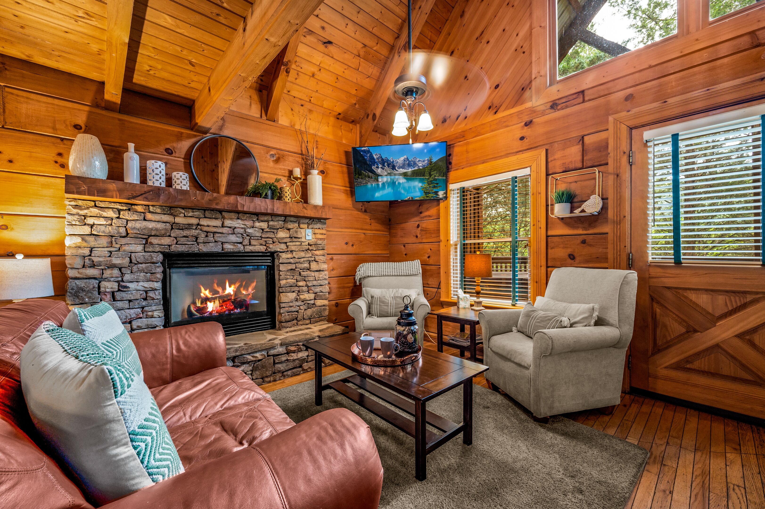 A cozy cabin interior with a stone fireplace, leather sofa, and two armchairs.