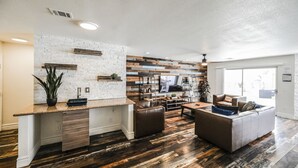 A modern living room with a large TV, leather sofas, and a kitchen area with a white brick backsplash and wooden accents. - SVS: Spacious 6BD Vegas Home | Private Pool, BBQ & Game Room | Sleeps 12 (Henderson)