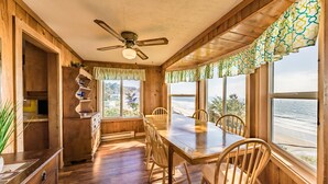 Comedor de madera con una mesa, sillas, ventilador de techo y ventanas grandes con cortinas.