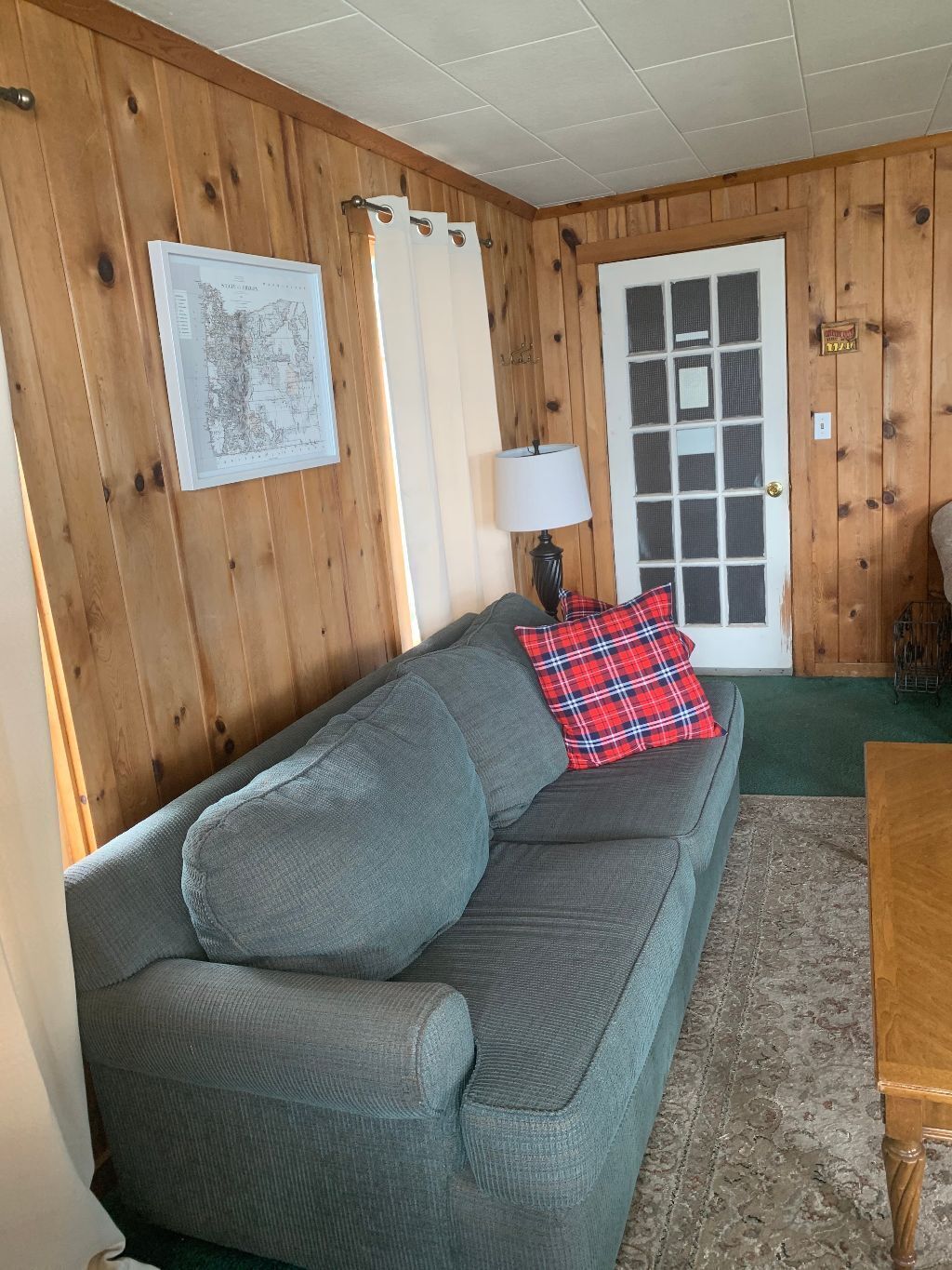 Ein gemütliches Zimmer mit einer holzverkleideten Wand, einem grauen Sofa mit kariertem Kissen, einer Lampe und einer Tür mit Fenster.