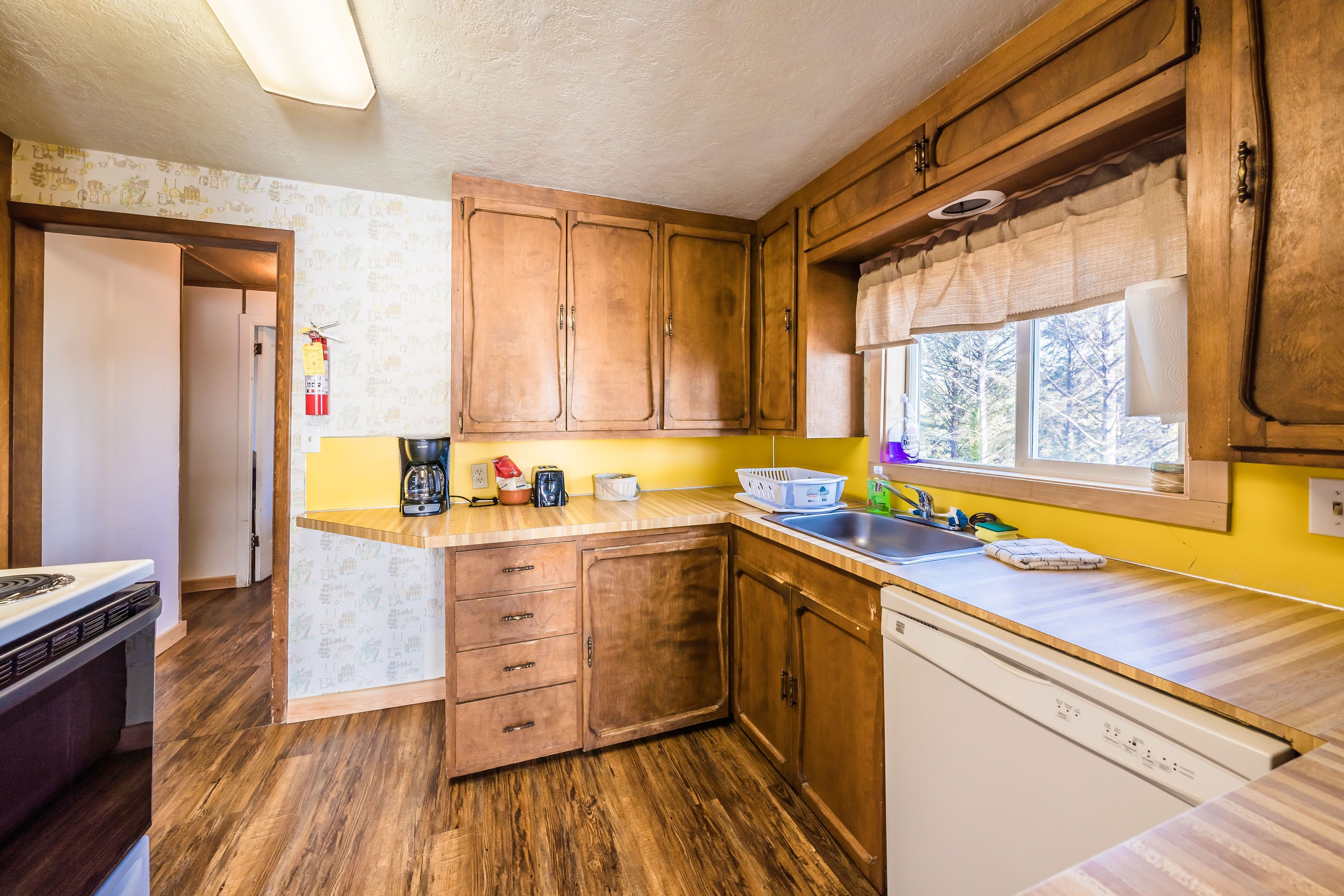 A compact kitchen with wooden cabinets, a built-in dishwasher, and a window with a view of trees.
