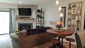 A modern living room with a stone fireplace, a flat-screen TV, a sofa, a round dining table with stools, and a decorative wall with building images. - 1 Bedroom, 1 bath townhouse in The Galena Territory. (Galena)