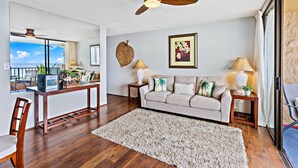 A living room with a wooden floor, a beige sofa, two side tables with lamps, a ceiling fan, and a large window with a view. - Open, spacious 1bd  with oceanfront views (Lahaina)