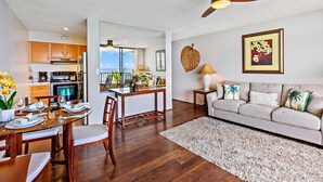 A modern living room with a sofa, a dining table set for four, and a kitchen area with wooden cabinets and a ceiling fan. - Open, spacious 1bd  with oceanfront views (Lahaina)