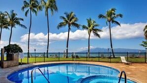 A swimming pool with a view of the ocean and mountains, surrounded by palm trees and a fence. - Open, spacious 1bd  with oceanfront views (Lahaina)