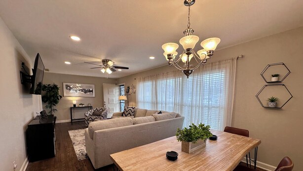 A dining area with a wooden table, brown leather chairs, and a chandelier. There is a ceiling fan and a sofa in the background.