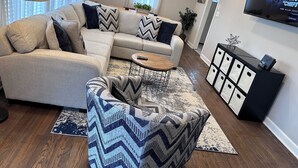 A living room with a sectional sofa, a round coffee table, and a patterned ottoman.