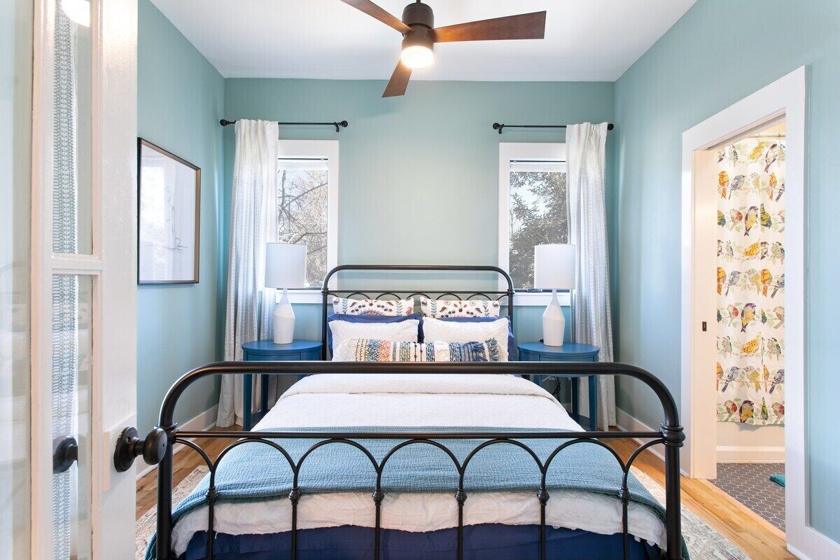 A bedroom with a bed, a ceiling fan, two windows with curtains, a door with a bird-patterned wallpaper, and a framed picture on the wall.