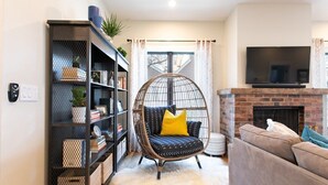 Ein Wohnzimmer mit schwarzem Rattanstuhl, einer Bücherwand, einem grauen Sofa und einem Kamin.