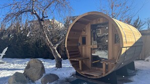 Eine hölzerne, fassförmige Konstruktion mit einer Sitzbank im Inneren, umgeben von Schnee und Steinen.