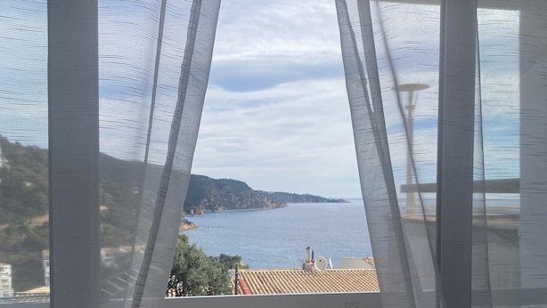 Un balcón con vista al mar y colinas distantes, cortinas entreabiertas y piso de baldosas.