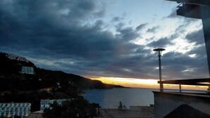 Vista costera con un atardecer sobre el agua, edificios a la izquierda y un farol a la derecha.