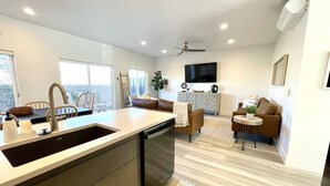 A modern kitchen with a large island, a dining area with a sofa and chairs, a flat-screen TV mounted on the wall, and a ceiling fan. - Trendy 5 BD/4.5 BA St. George Townhome at The Vue (St. George)