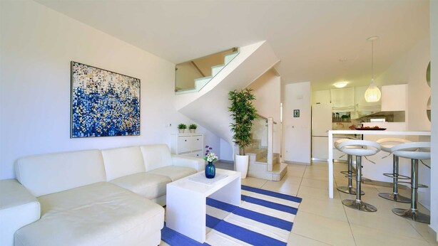 Ein modernes Wohnzimmer mit einem weißen Sofa, einem Couchtisch und einem blau-weiß gestreiften Teppich. Eine Treppe führt auf eine erhöhte Ebene, es gibt eine Küchenzeile mit weißen Schränken sowie einen Essbereich mit Barhocker.