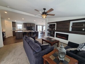 Een moderne woonkamer met een bank, salontafel en open haard. Achtergrond toont een keuken met een eethoek.
