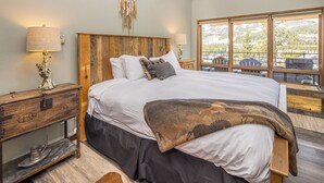 Un dormitorio con una cama grande, cabecera de madera, mesitas de noche con lámparas, un atrapasueños y vistas al exterior a través de amplios ventanales.