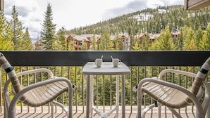 Een balkon met twee witte metalen stoelen en een kleine tafel, met uitzicht op een bebost berglandschap met gebouwen.